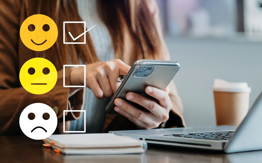 Eine Frau sitzt am Schreibtisch mit einem Smartphne in der Hand, auf dem Foto sind 3 Smilies von gut bis schlecht, wobei gut angehakt ist